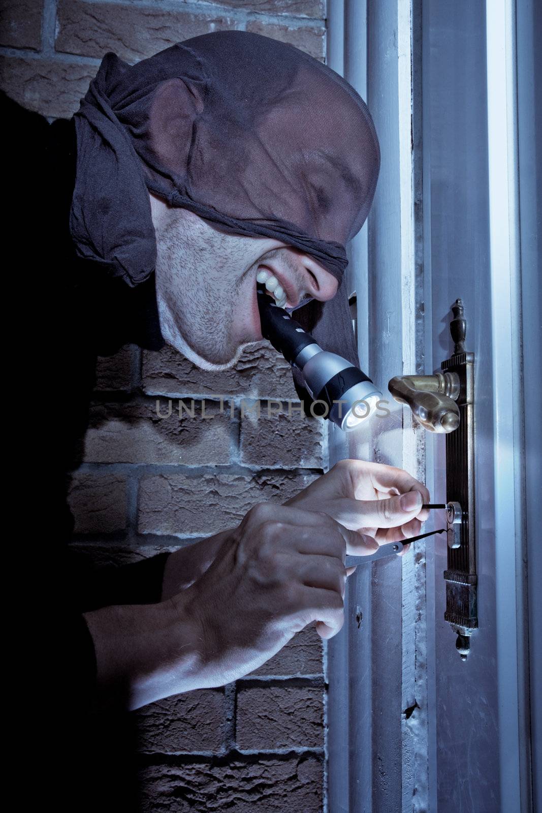 Masked burglar holding a flashlight in his mouth, picking the lock of a door.