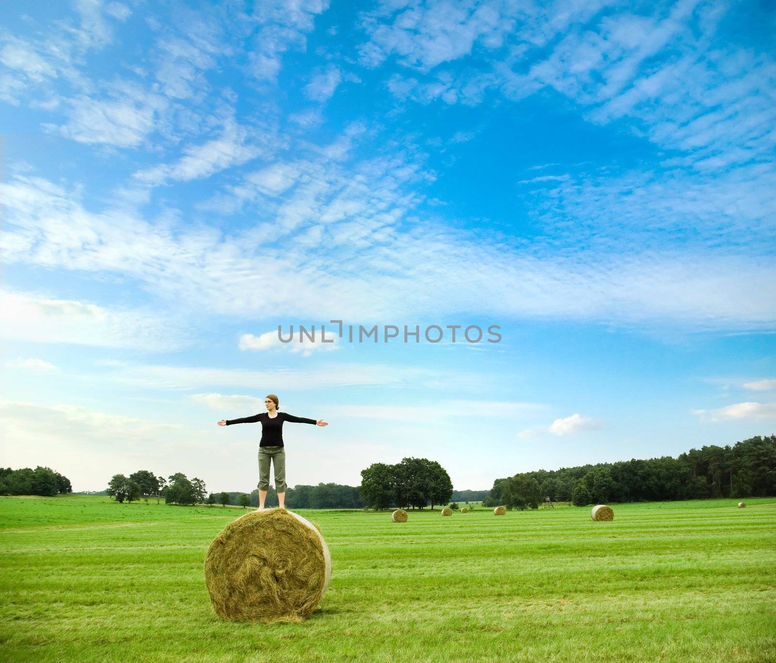 woman balancing on top of a hay bale on a meadow, infront of  a blue sky