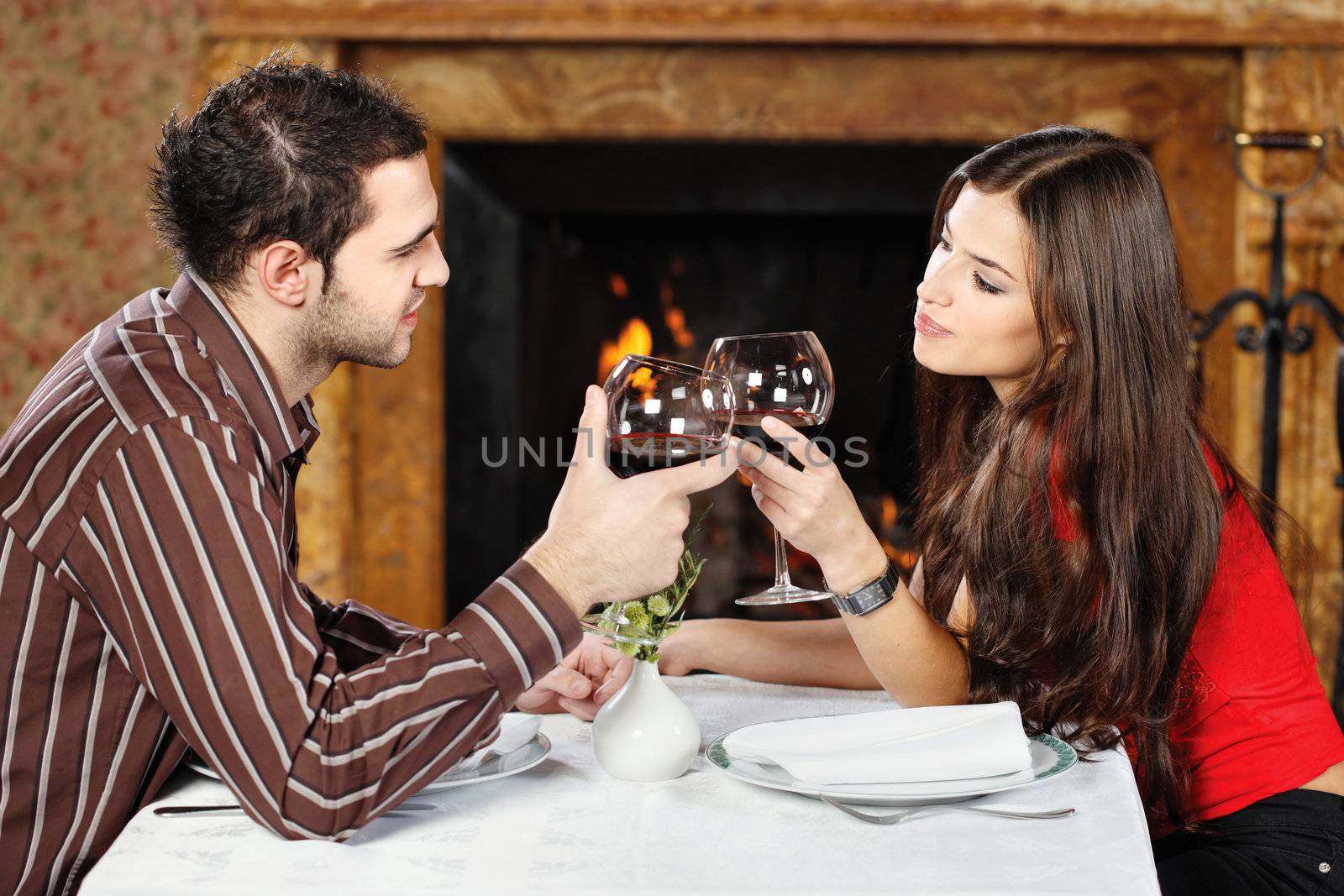 Young couple in love near fireplace, holding glasses of wine and cheers