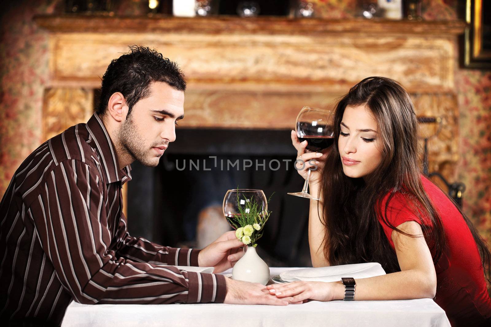 She hold his hand while they sitting at table and drinking wine near fireplace, focus on male