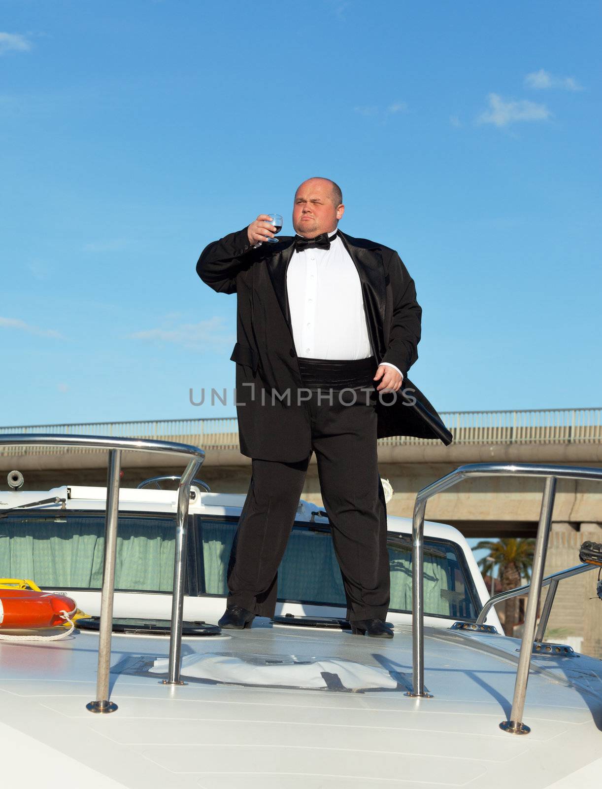 Overweight man in tuxedo standing on the deck of a luxury pleasure boat with glass red wine