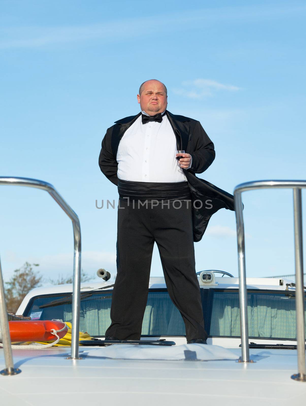 Overweight man in tuxedo standing on the deck of a luxury pleasure boat with glass red wine