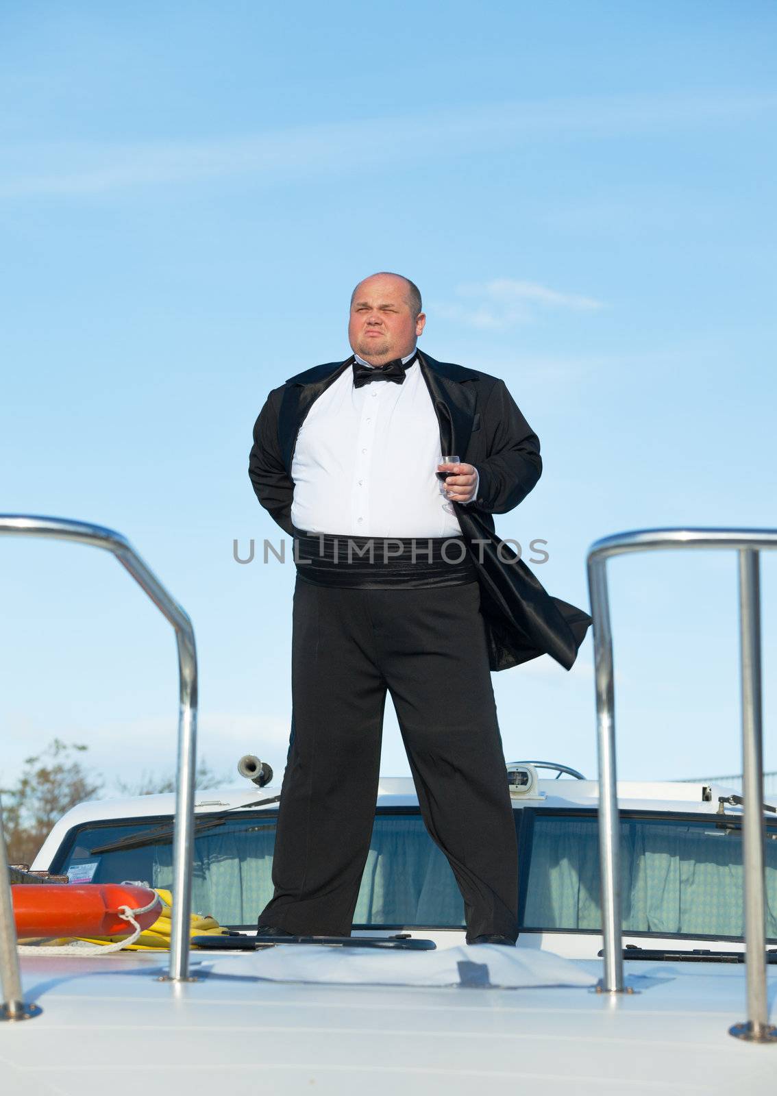 Overweight man in tuxedo standing on the deck of a luxury pleasure boat with glass red wine