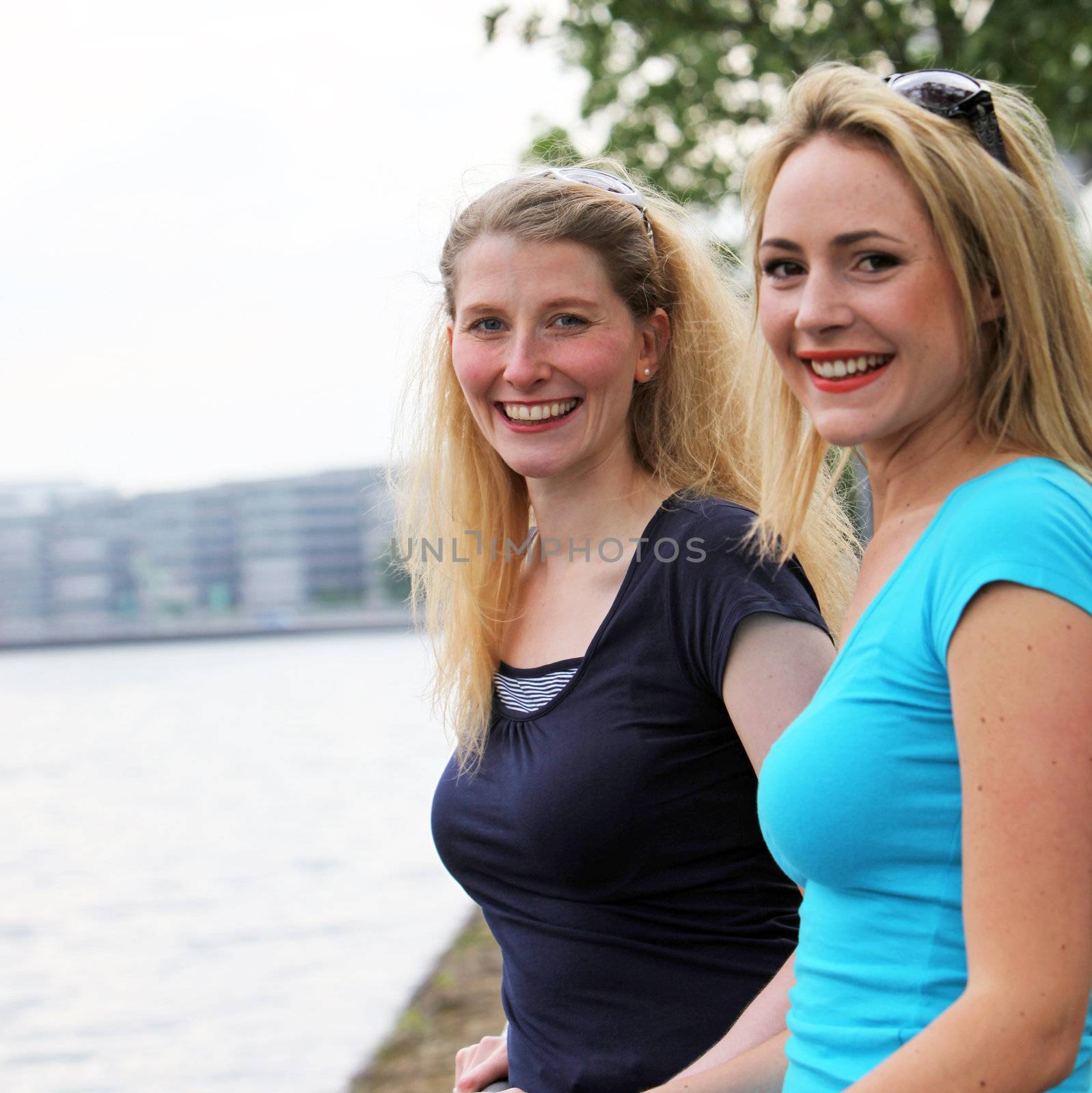 Two smiling happy women standing side by side overlooking a river as they enjoy an afternoon relaxing in each others company 