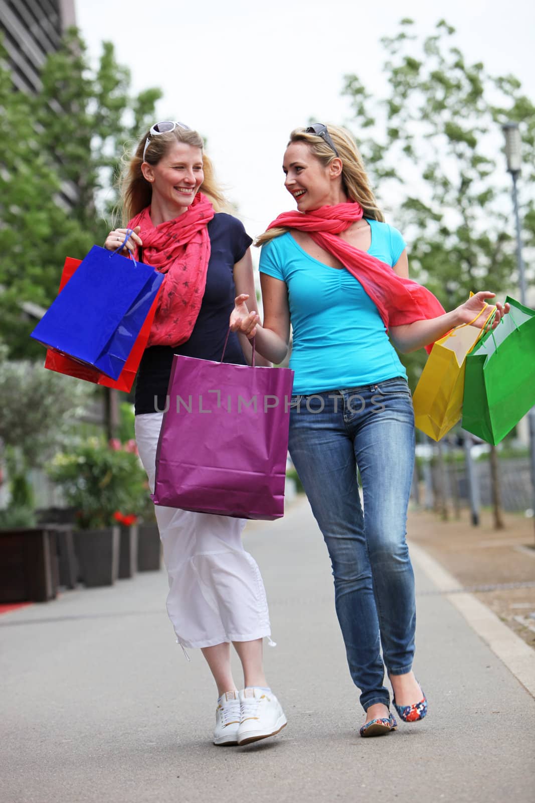 Two female friends on a successful shopping trip walking along the sidewalk chatting happily Two friends on a successful shopping trip walking along the sidewalk chatting happily 