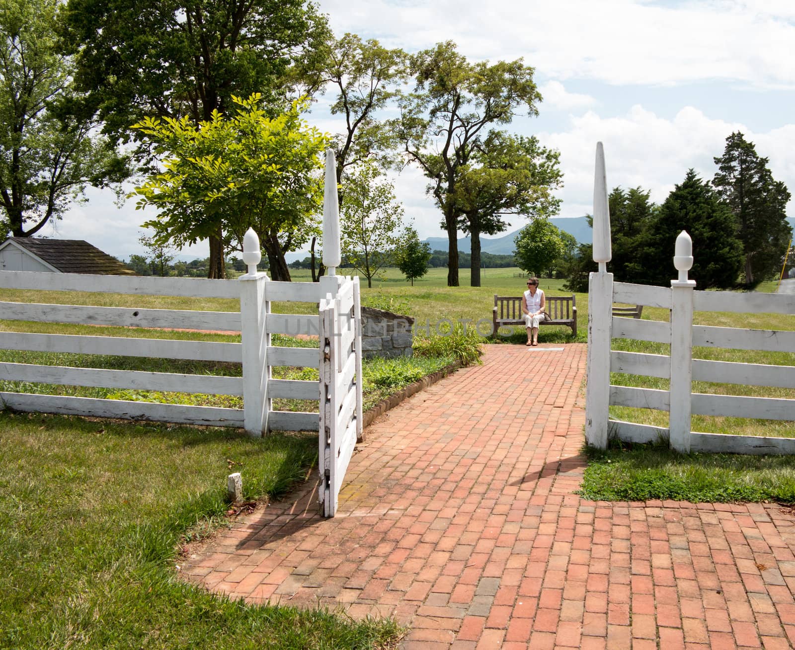 Senior woman sitting on bench on brick path through white picket gate