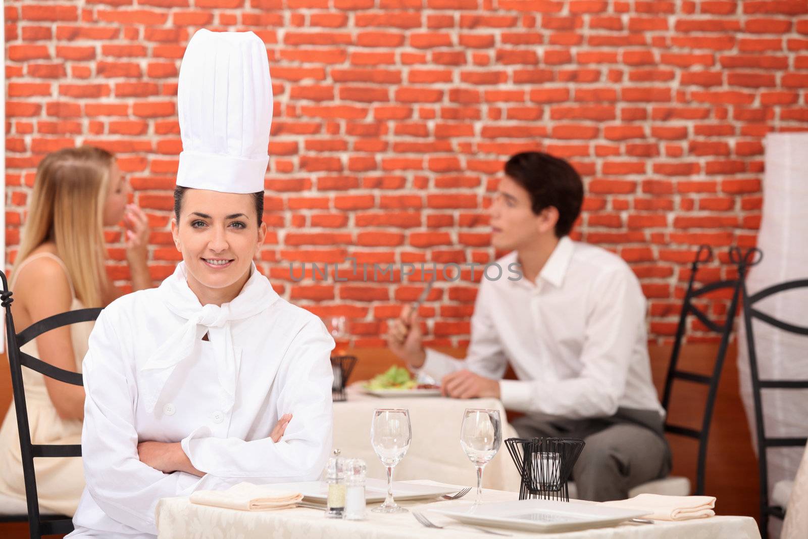 female chef posing in restaurant with couple dining in background