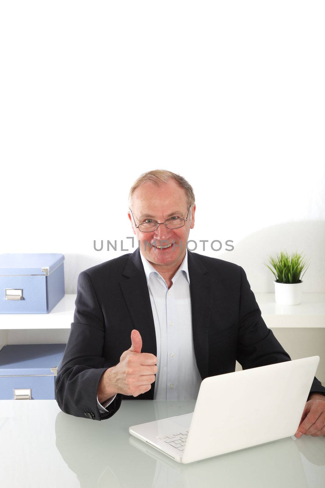 Senior businessmanseated at his desk with his laptop giving a thumbs up gesture of agreement 