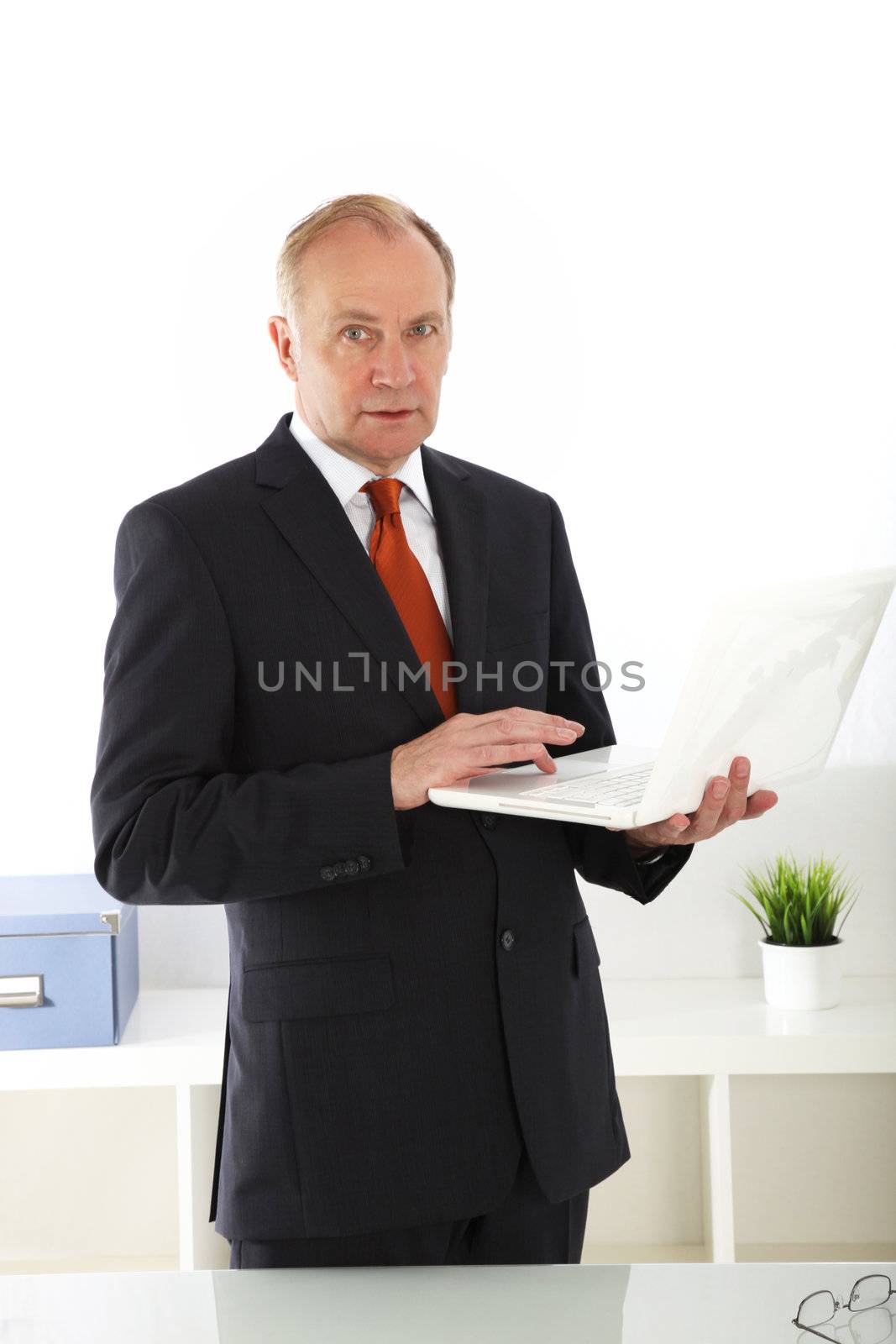 Senior stylish business executive standing working on his laptop which is balanced on his arm as he waits for the start of a meeting 