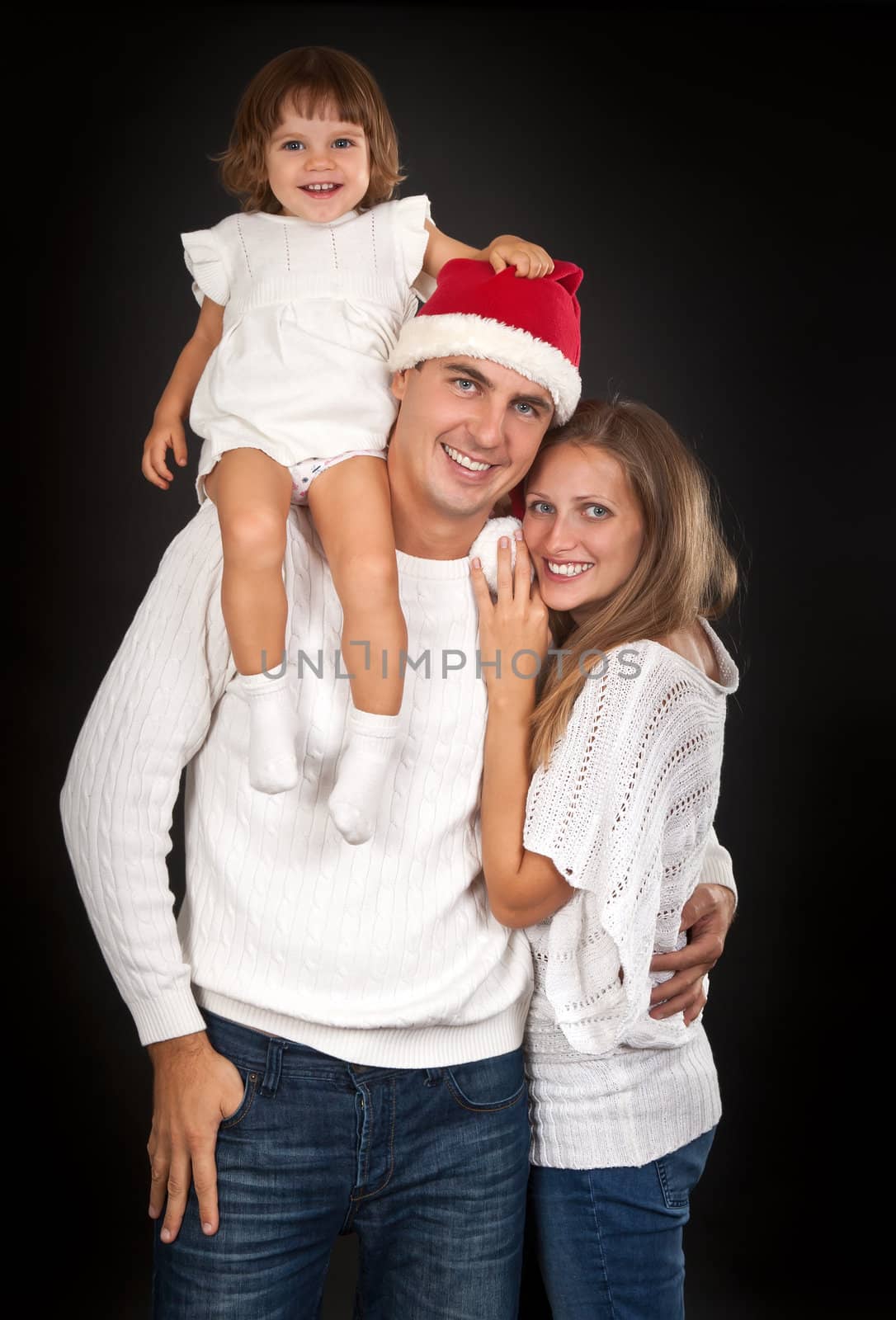 Father in santa hat holding little girl on his shoulder and hugged his wife in the studio against a dark background