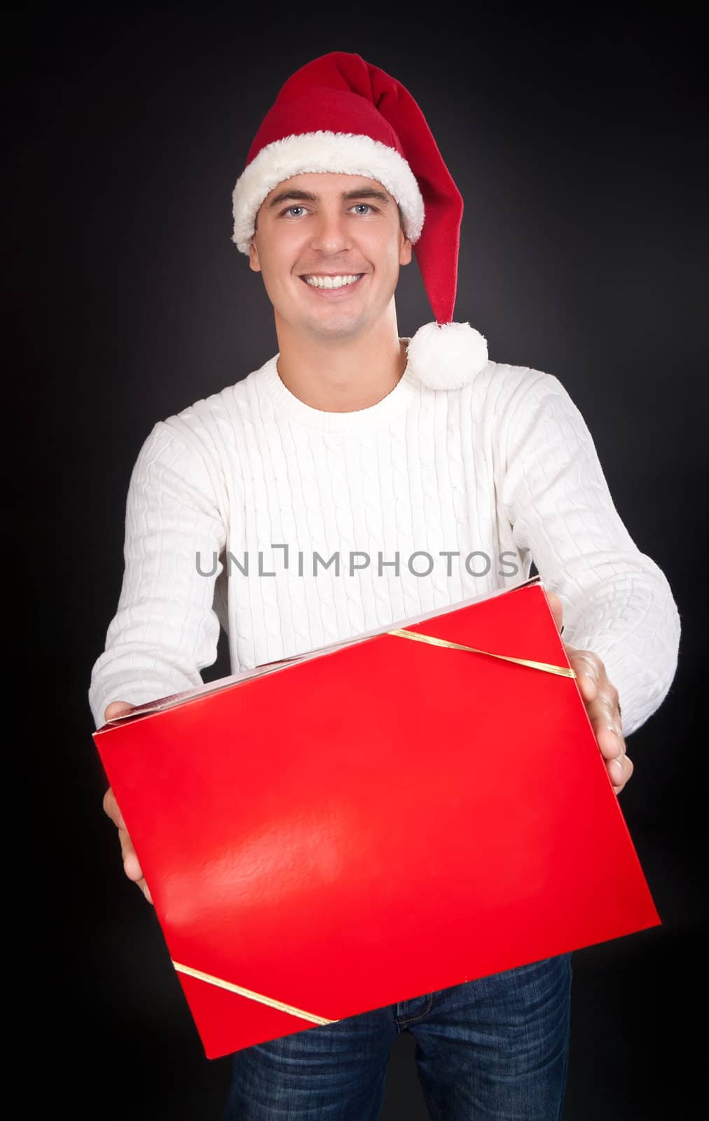 Smiling man in Santa hat gives a red box in the studio against a dark background