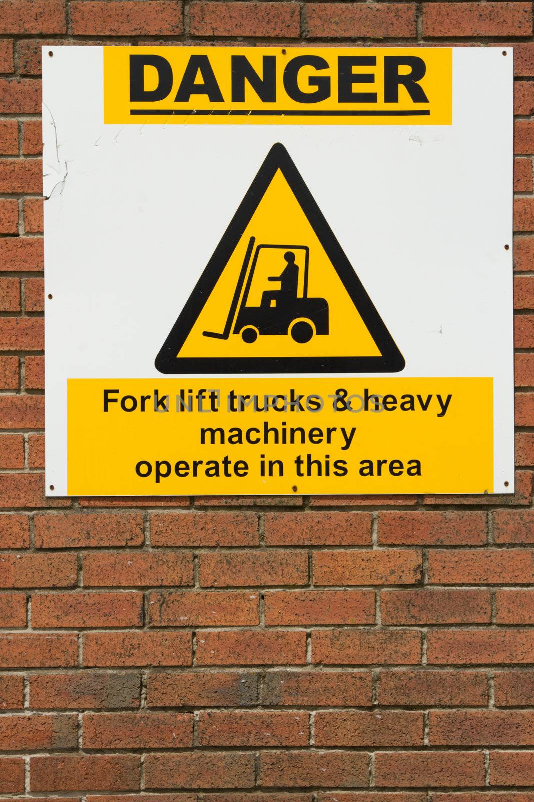 A white sign on a red brick wall with the symbol for machinery and the words 'DANGER' in bold and the words 'fork lift trucks & heavy machinery operate in this area'.