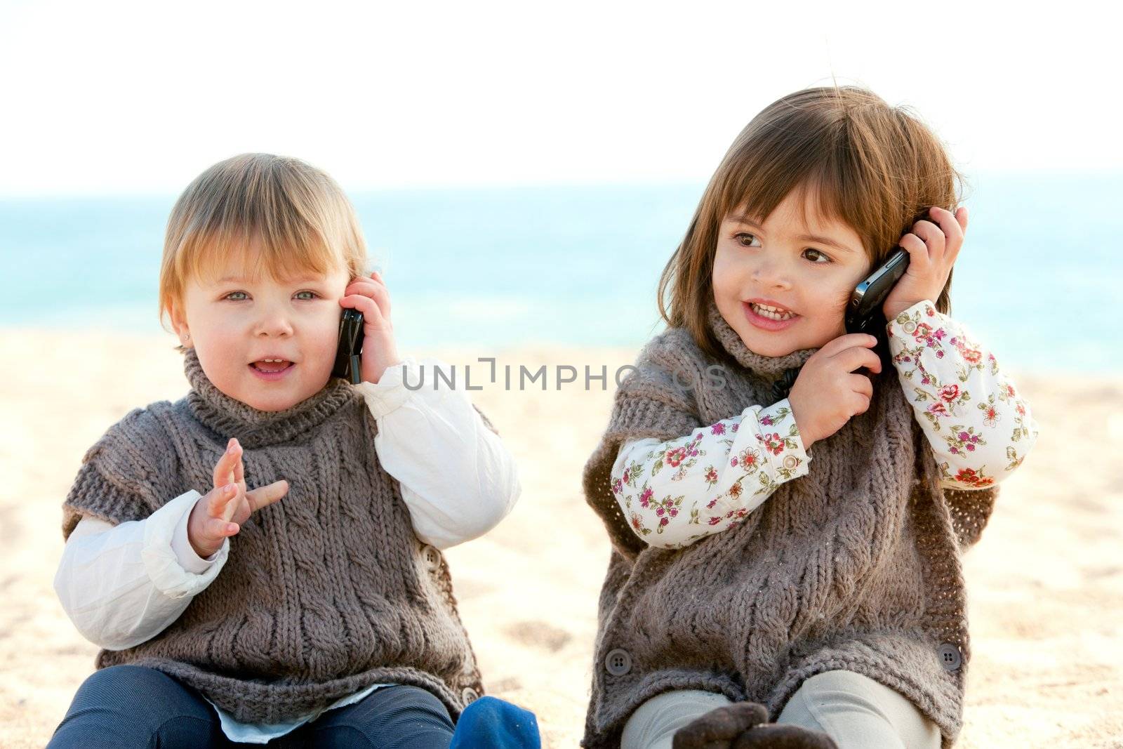 Portrait of two cute little girls on beach having conversation on mobile phones.