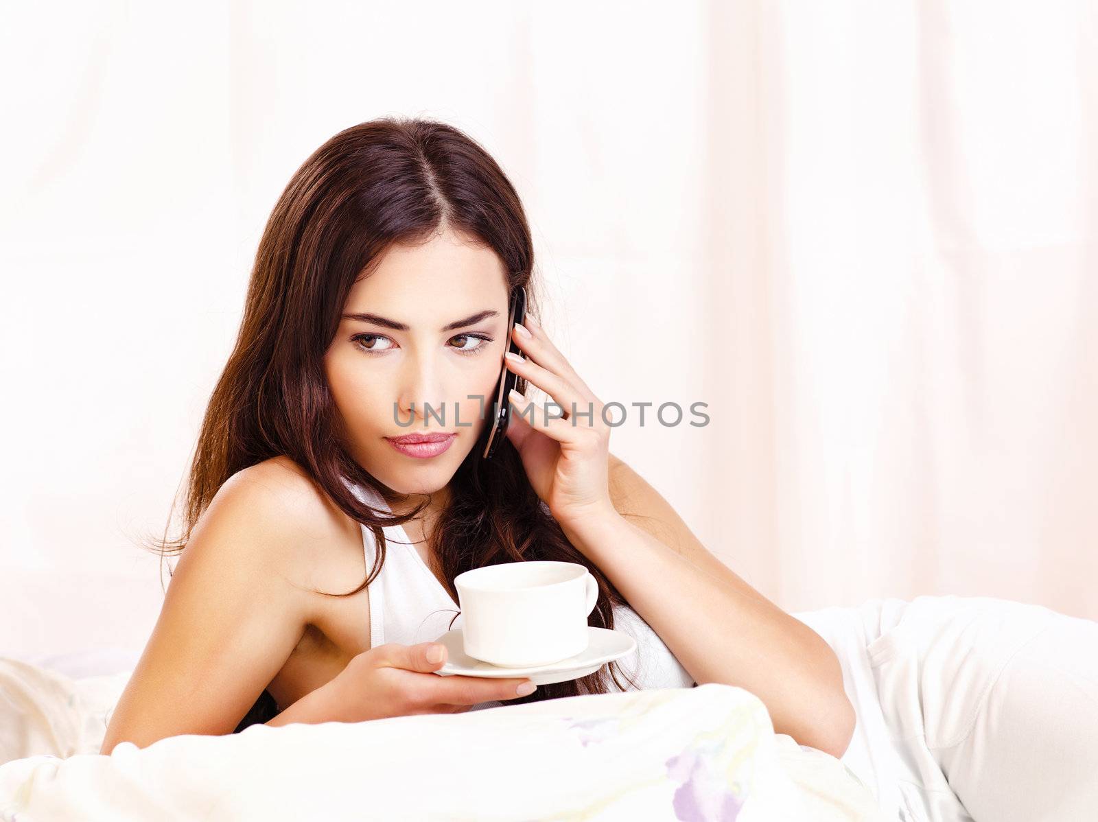 Pretty woman making phone call and holding cup of coffee in bed
