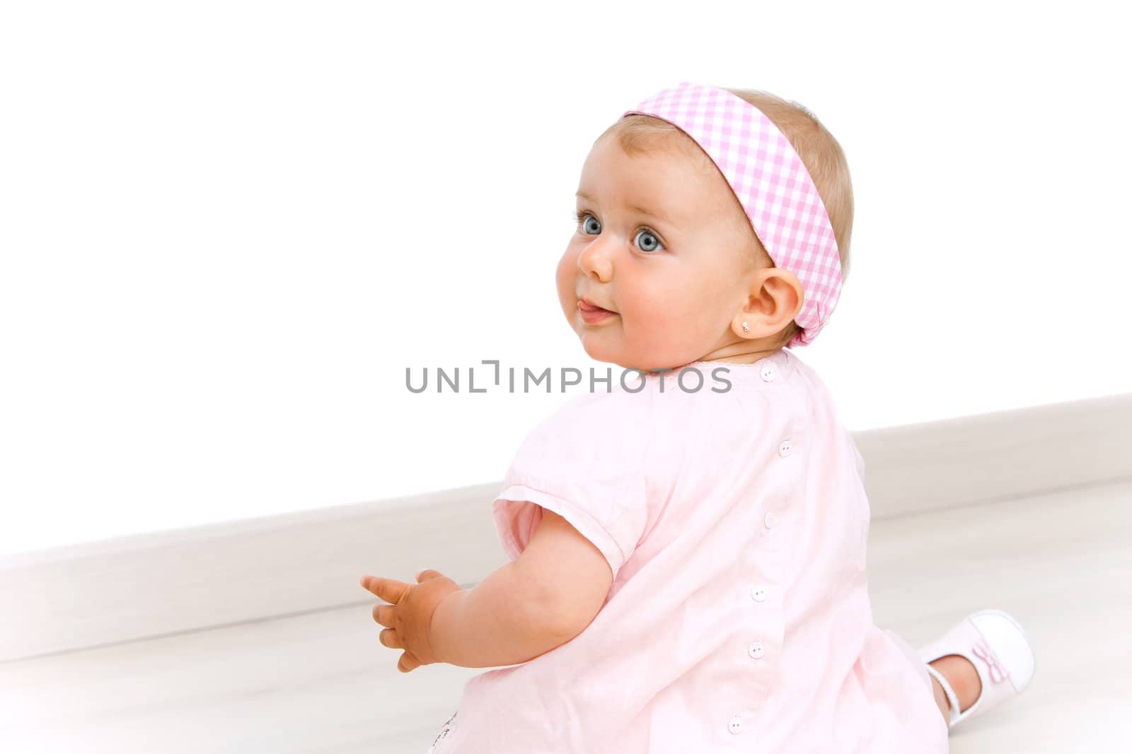 Blue eyed baby girl wearing a pink dress and pink head band sitting on the floor.