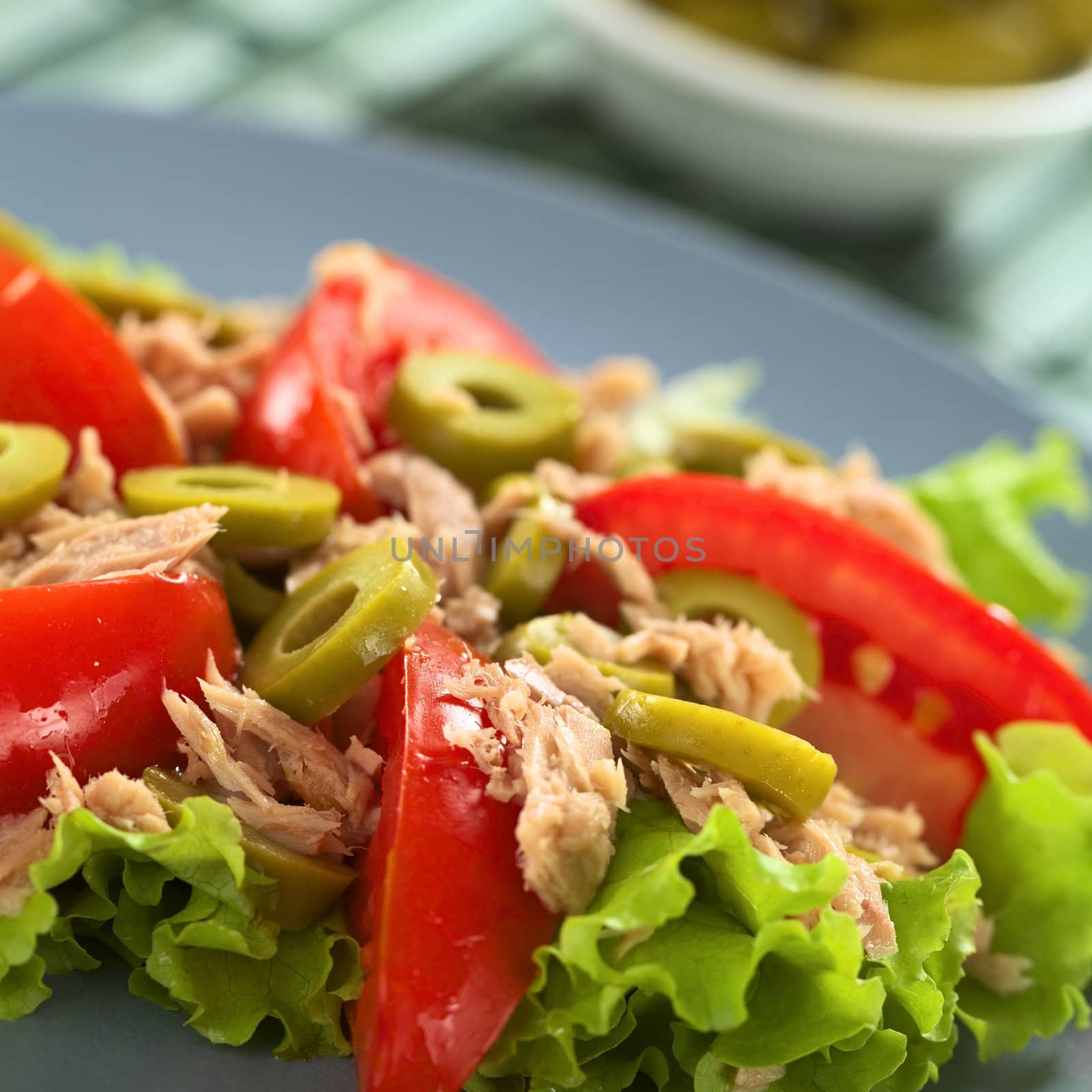 Fresh tuna, tomato and green olive salad served on lettuce leaf on blue plate with olives in the back (Selective Focus, Focus on the front of the olive in the middle of the salad) 