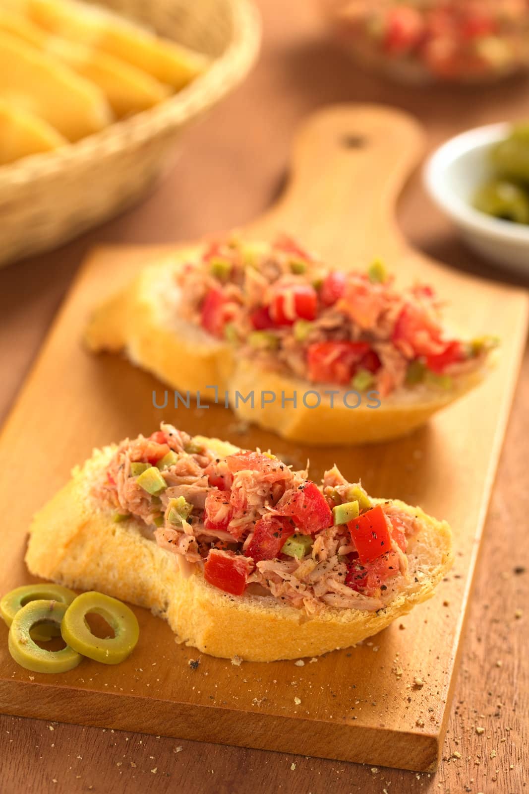 Slice of baguette with fresh tuna, green olive and tomato spread seasoned with ground pepper on wooden board (Selective Focus, Focus on the front of the spread on the first bread)