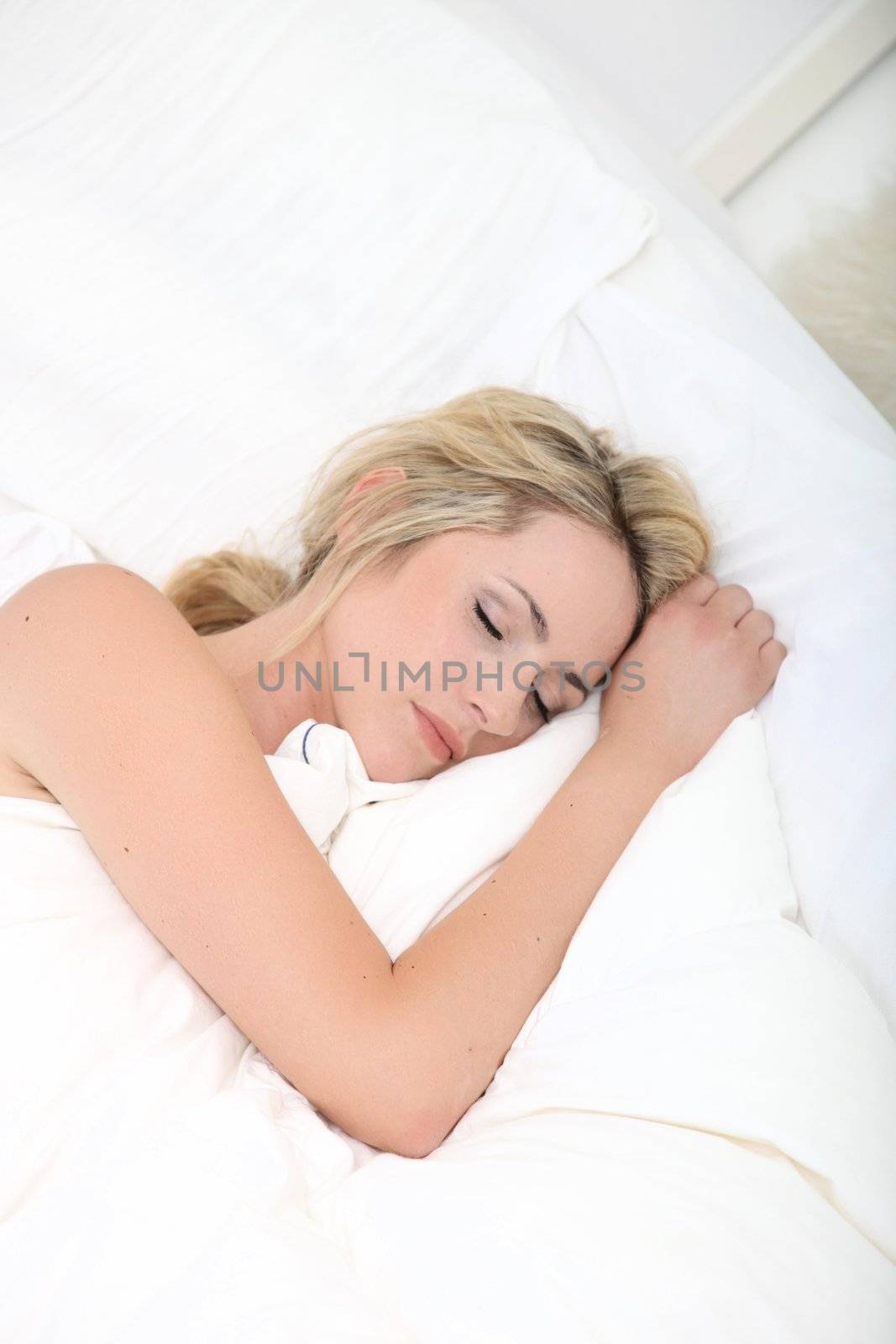 Natural view of a young blonde woman sleeping on white bedclothes with her arm out above the duvet as she tries to overcome an illness