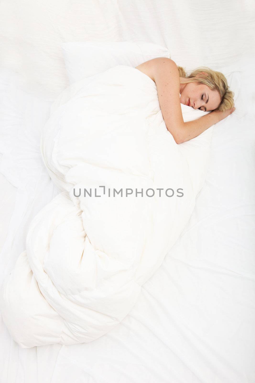 Overhead view of a sick woman curled up in a large white bed asleep under her duvet 