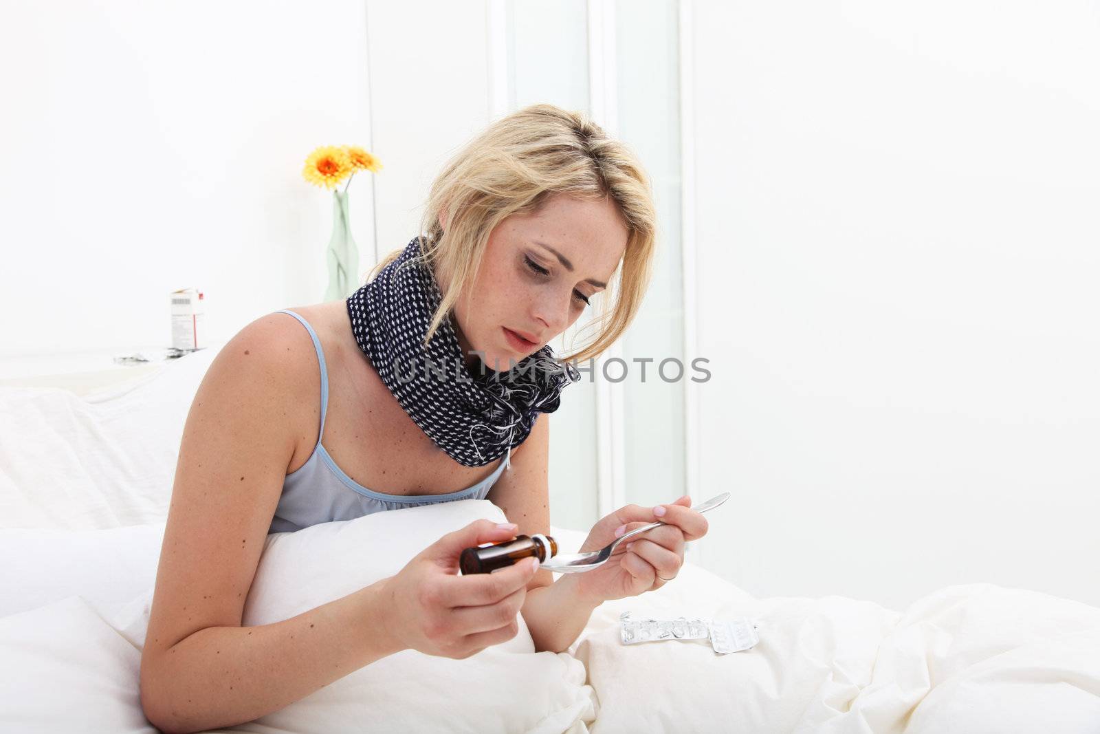 Sick woman sitting up in bed with a scarf wrapped around her neck pouring a dose of medicine into a spoon as she takes her medication 