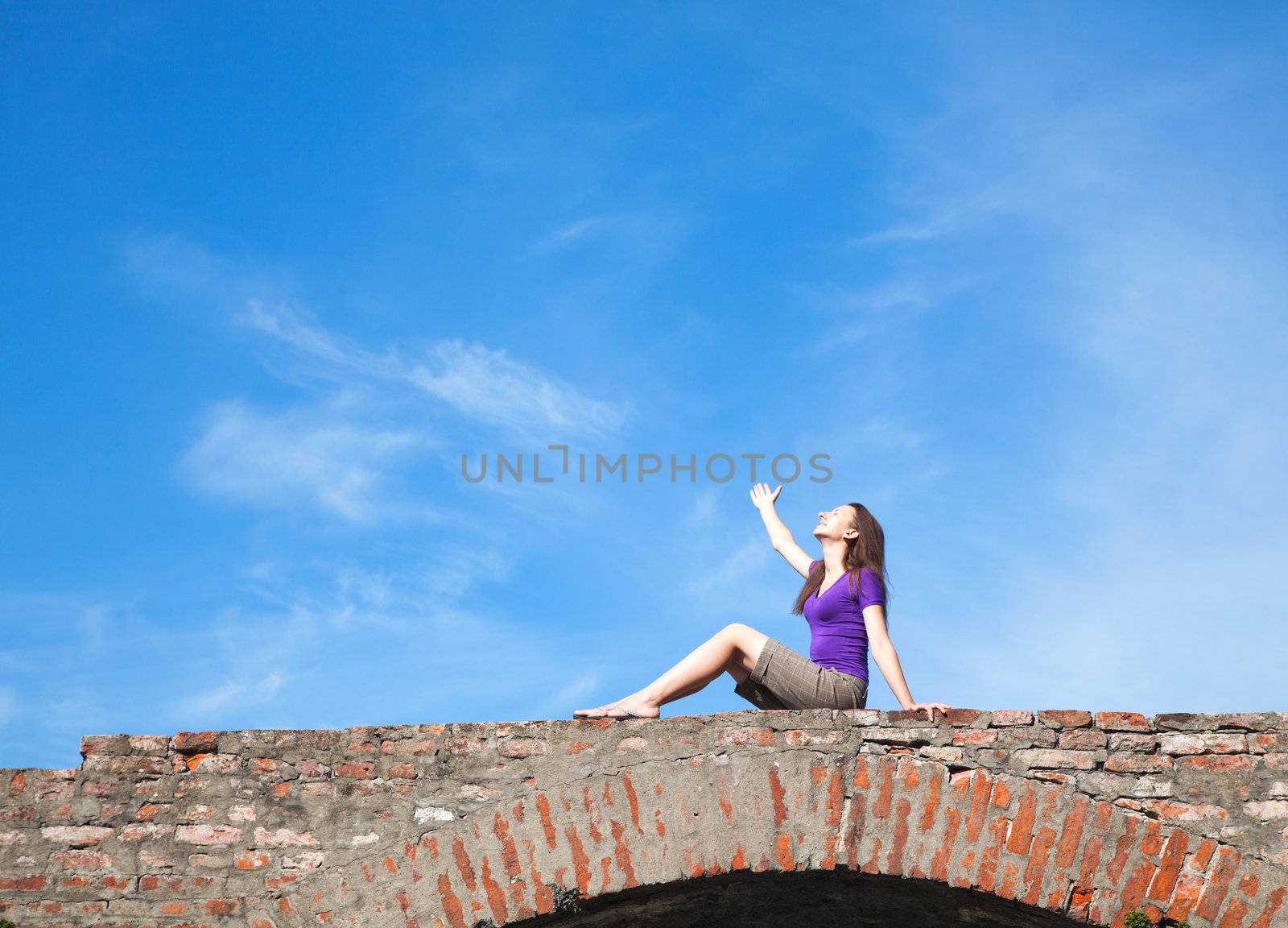 Young woman sitting with raised hand against blue sky