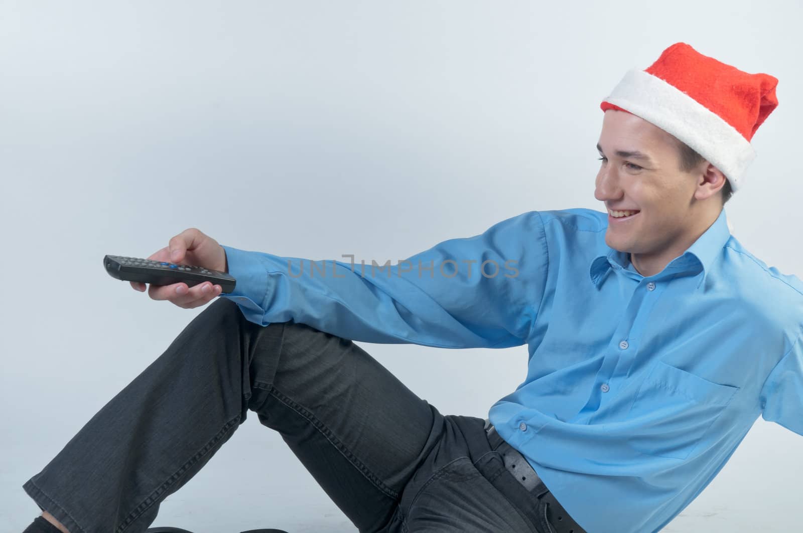 The young and handsome man in a red cap and a festive blue shirt on a white background, watching Christmas TV holding a TV remote