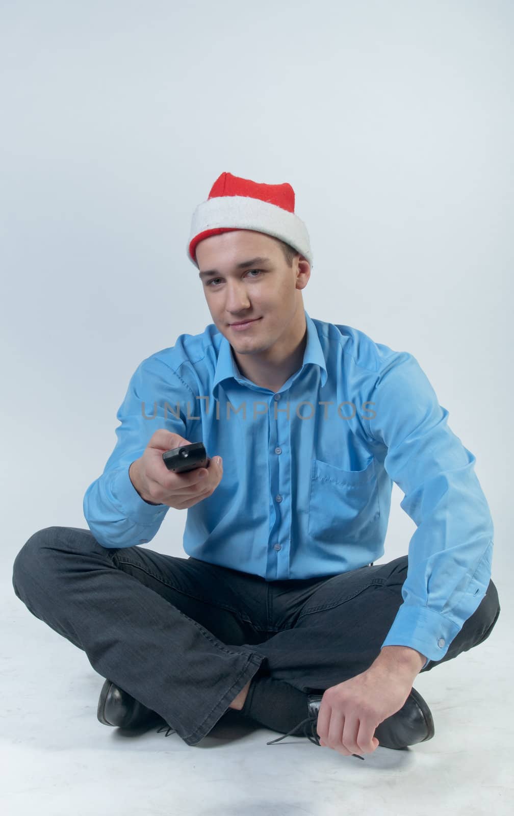 The young and handsome man in a red cap and a festive blue shirt on a white background, watching Christmas TV holding a TV remote