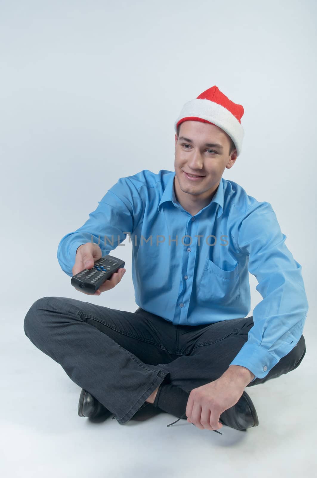 The young and handsome man in a red cap and a festive blue shirt on a white background, watching Christmas TV holding a TV remote