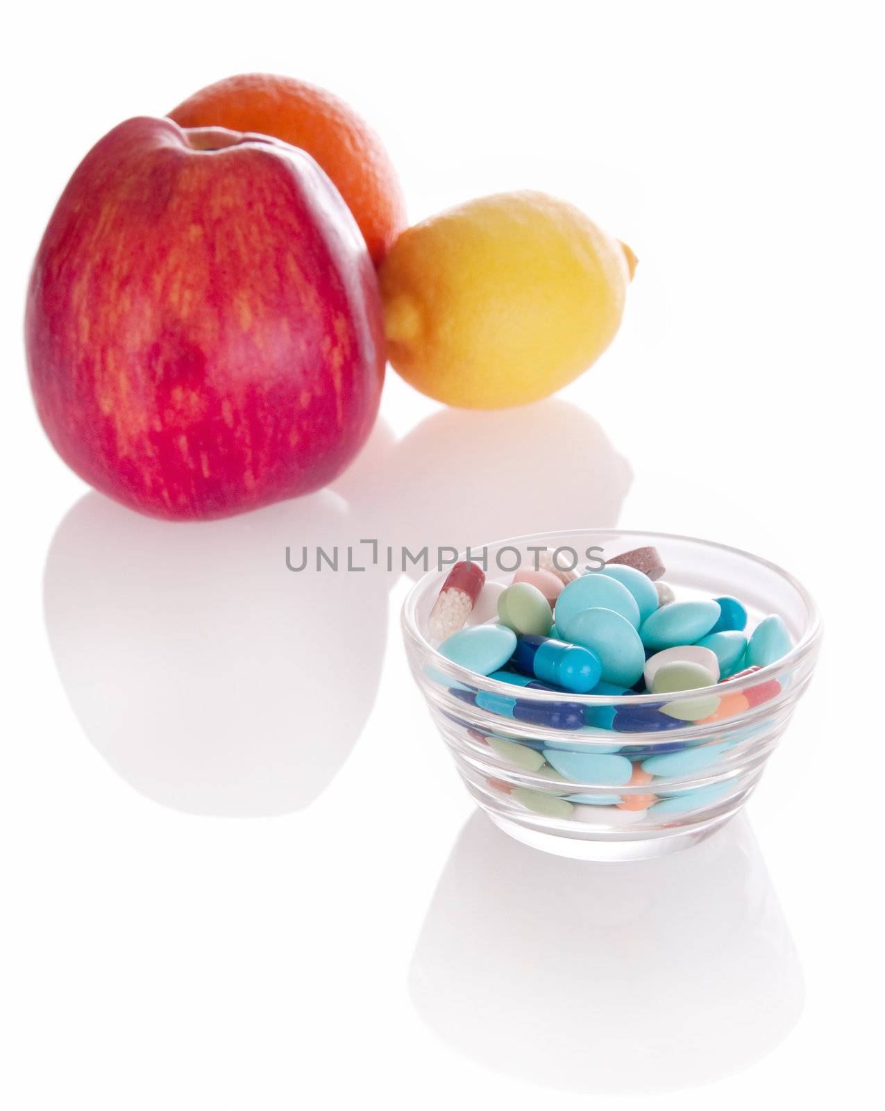 Concept of vitamin medicine with different pills in glass bowl and defocused fruits on background with soft shadows