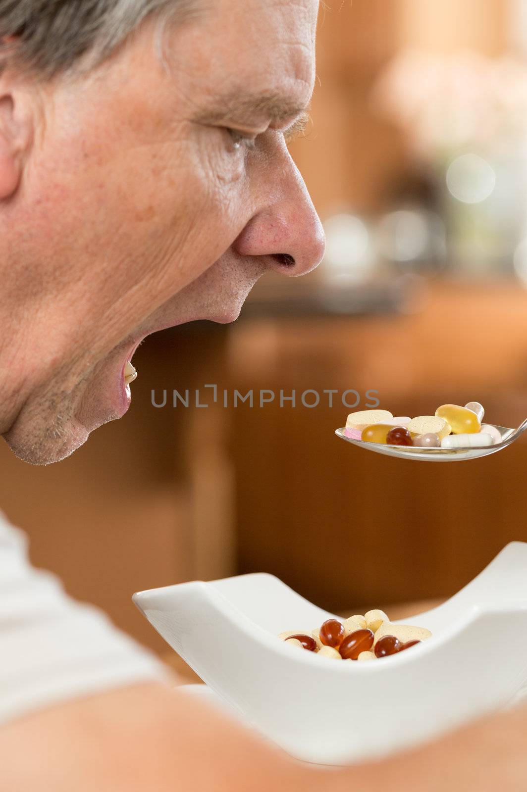 Senior caucasian male eating a spoonful of vitamin tablets for breakfast in kitchen