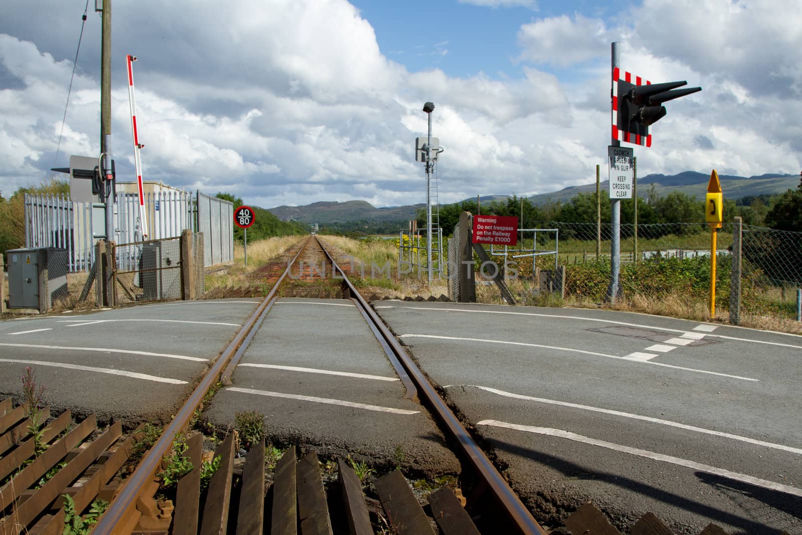 A rail line stretches into the distance with a tarmac road level crossing with safety markings, signs, lights and barriers.