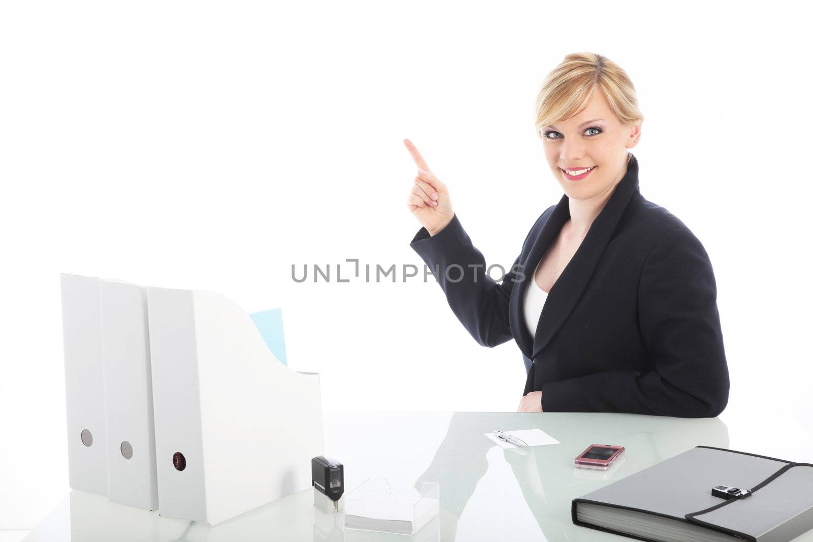 Smiling professional businesswoman seated at her white glass topped desk pointing to blank copy space above and to the side 