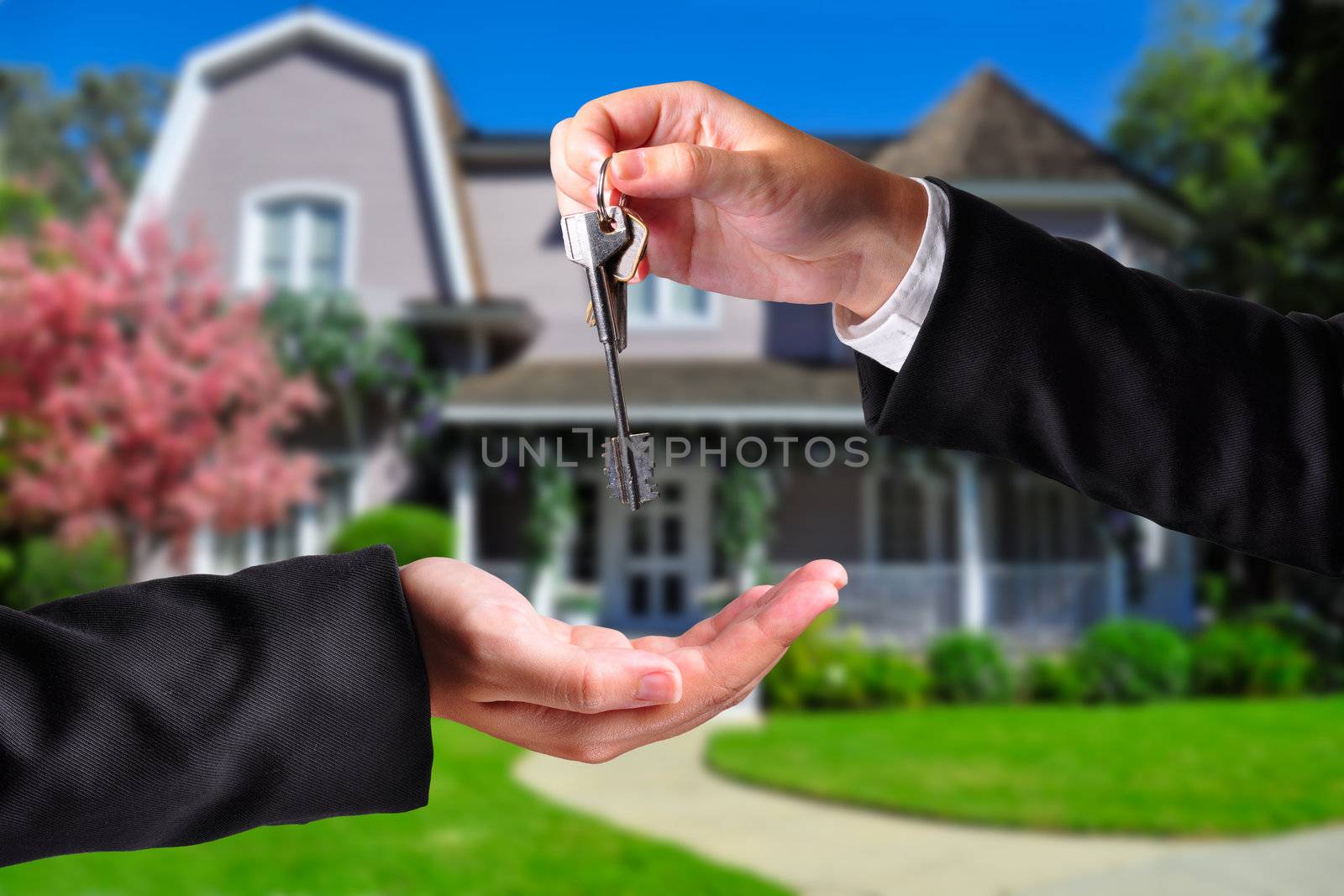 A hand giving a key to another hand. Both persons in suits and a house in the background.