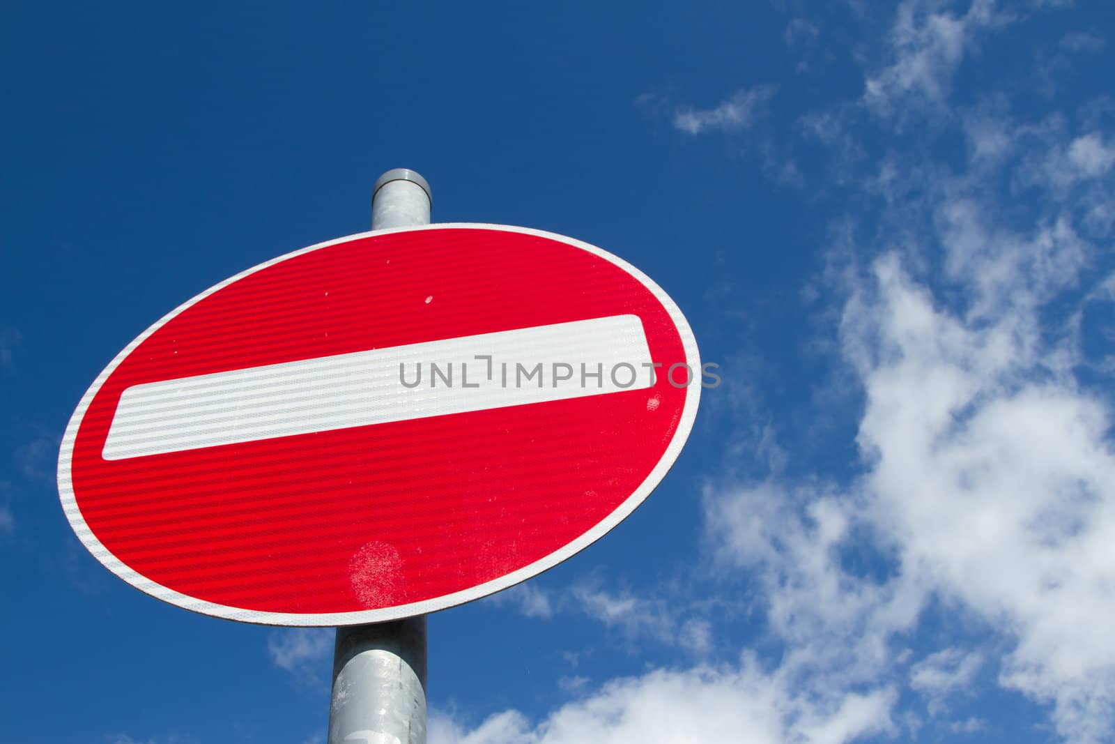 A no entry sign with a red circle and white bar on a metal post against a blue sky with cloud.