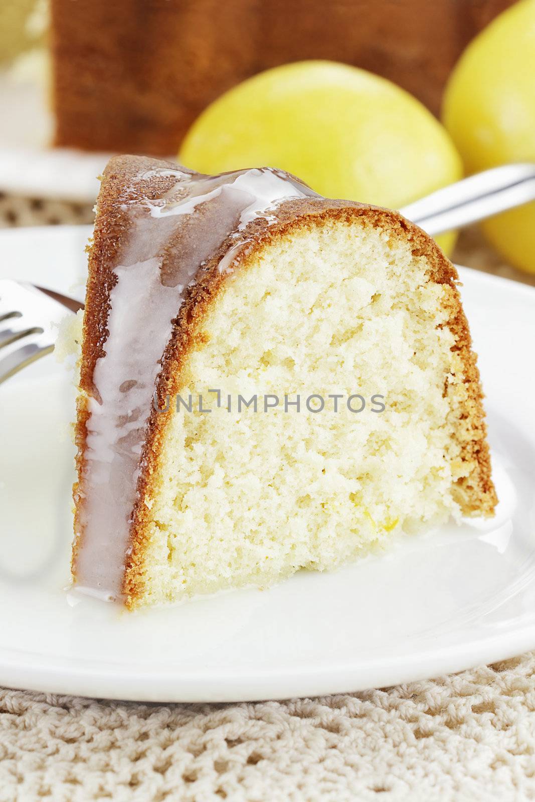 Slice of moist lemon bundt cake with lemons and cake in background. Shallow depth of field.