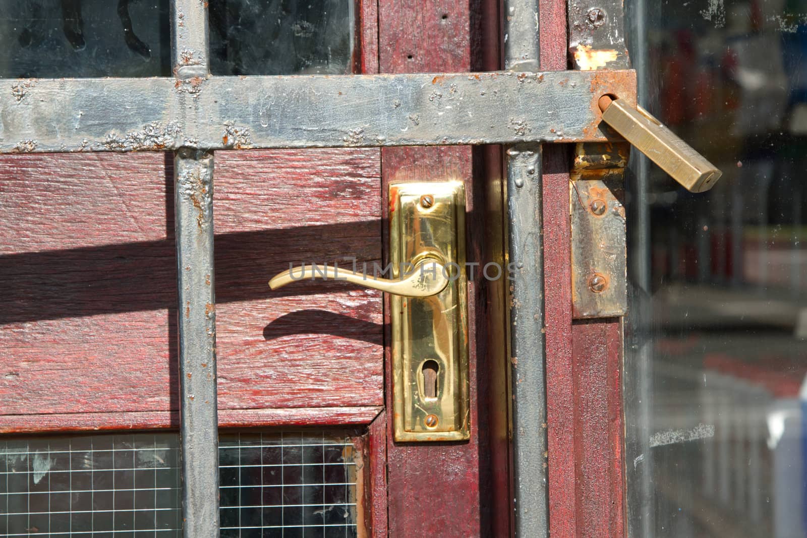 A wooden door with brass handle and keyhole protected by a metal grill secured with a metal padlock.