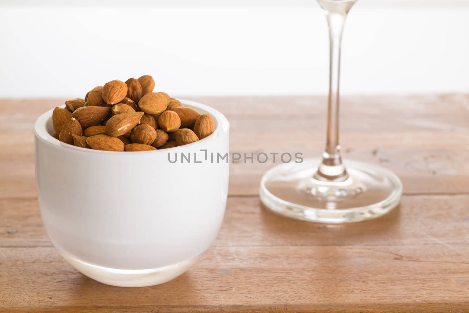 Raw almond nuts in white glass bowl on old wooden table with stem of wine glass