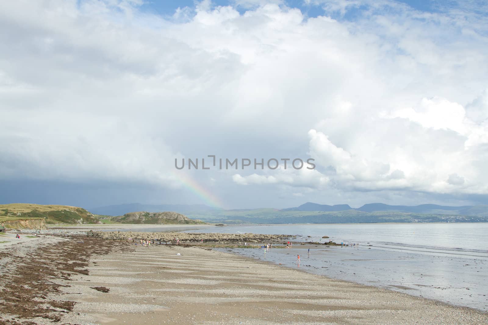 People on a sand and pebble beach in the sunshine with dark clouds over mountains in the distance with a rainbow.