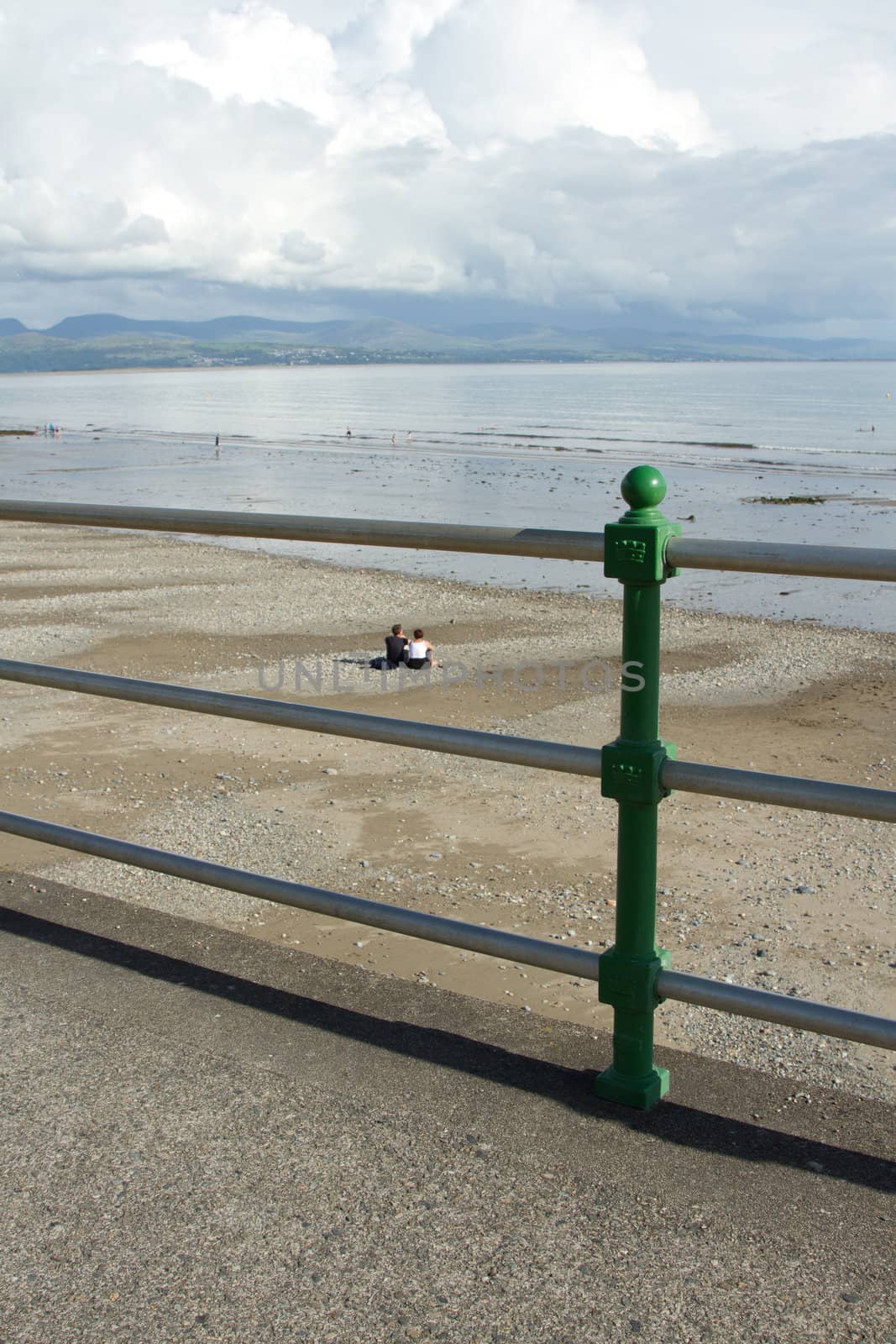 A set of railings over a sand and pebble beach with a couple sitting looking at clouds over the mountains in the distance.