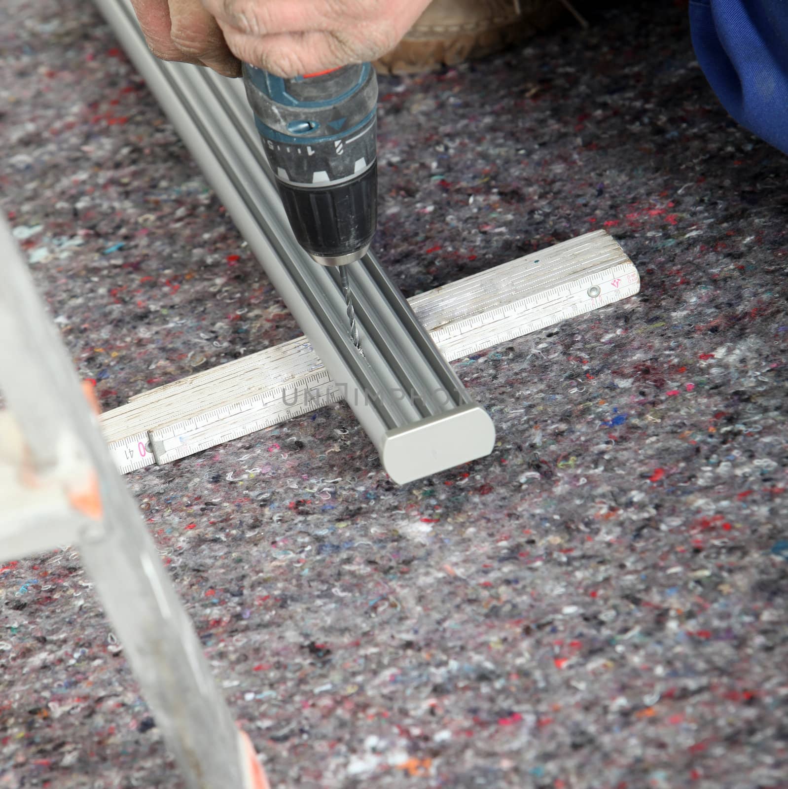 An artisan uses a portable cordless drill to drill a hole in a length of aluminium track supported at ground level.