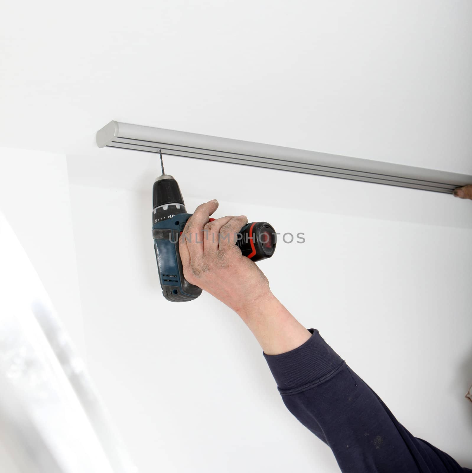 The arm of an artisan using a portable cordless drill to fit a length of aluminium curtain track to the ceiling
