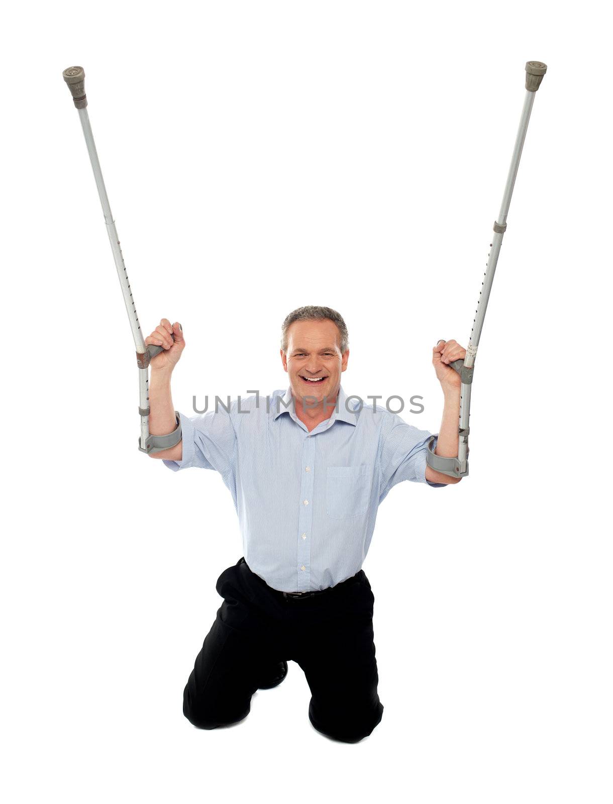 Cheerful senior man holding crutches upwards seated on floor, studio shot
