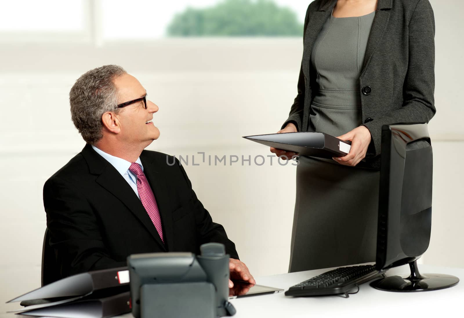 Smiling aged male boss looking at secretary while she presents business file to him