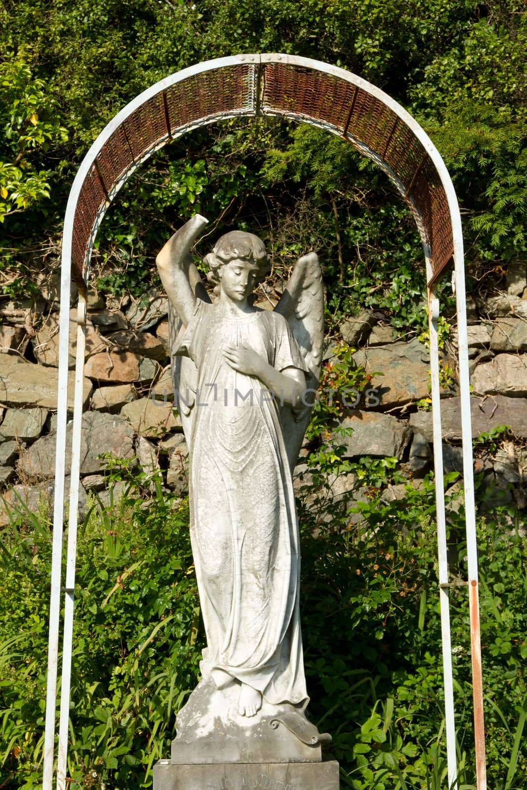 A stone carve statue of an angel, old and damaged under a metal frame with plants and a stone wall in the background.