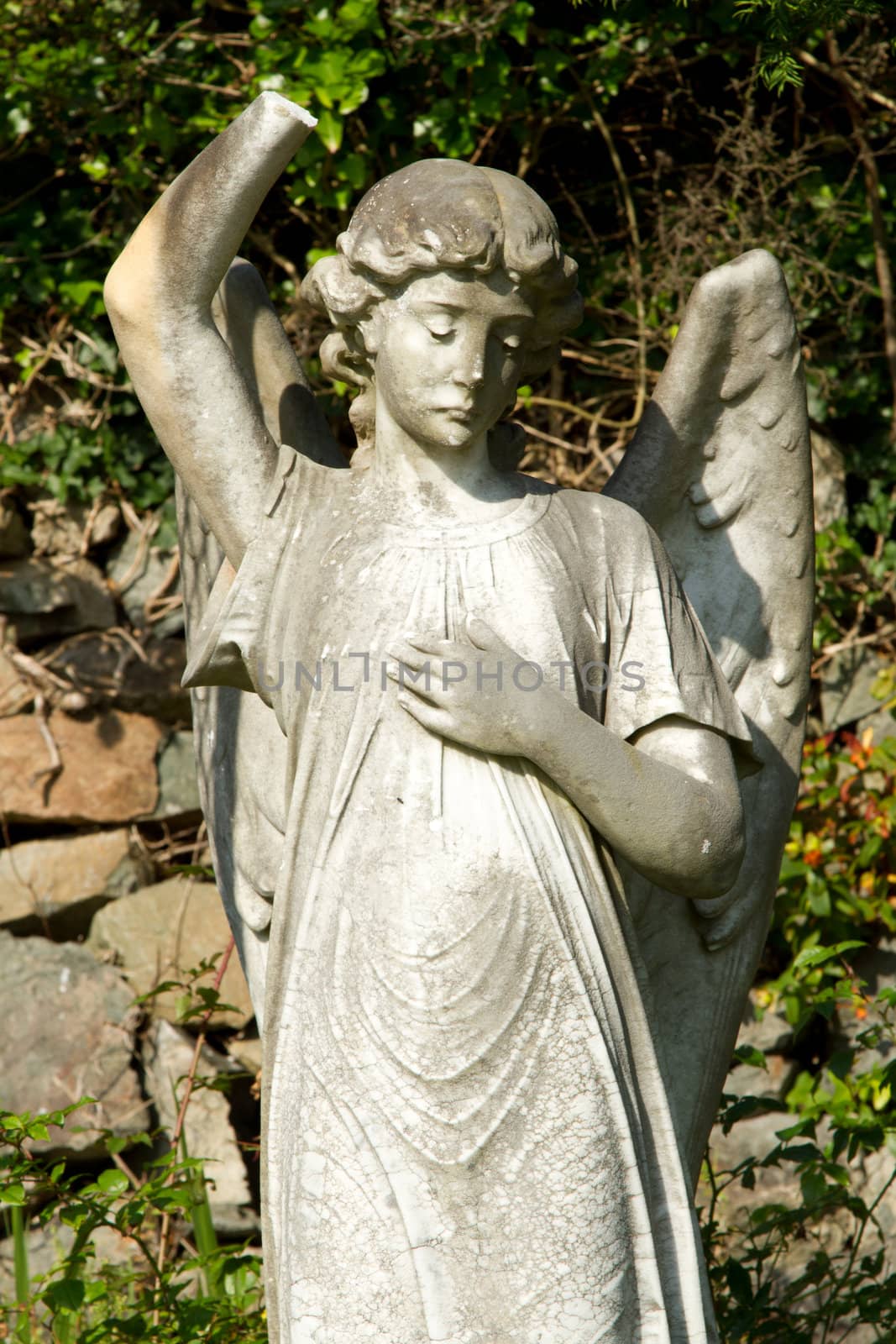 A stone carve statue of an angel, old, dirty and damaged with plants and a stone wall in the background.