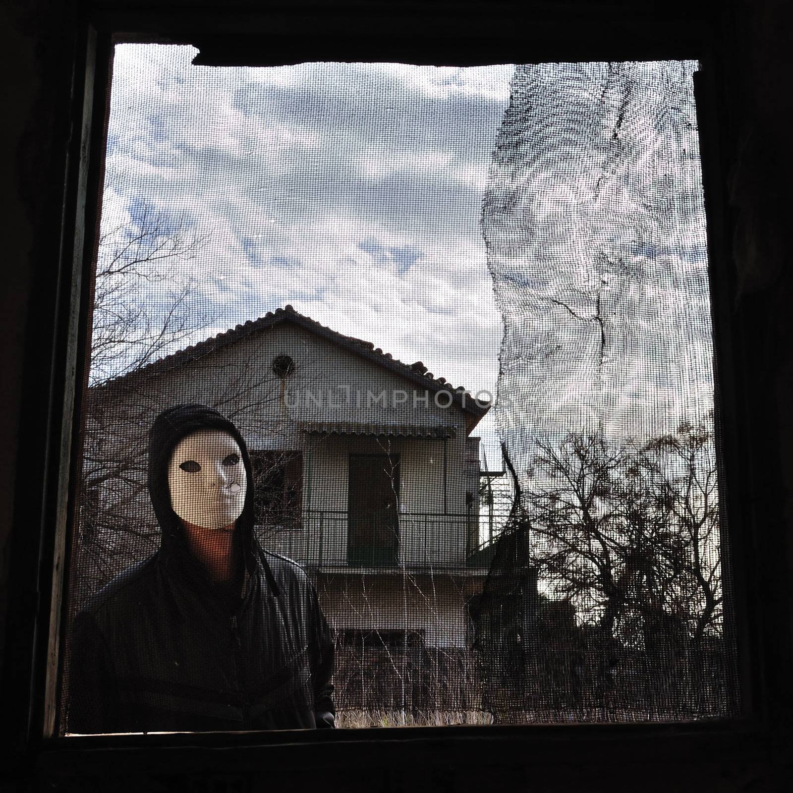 Hooded figure behind threaded window of derelict house. Masked maniac looking at you the viewer!