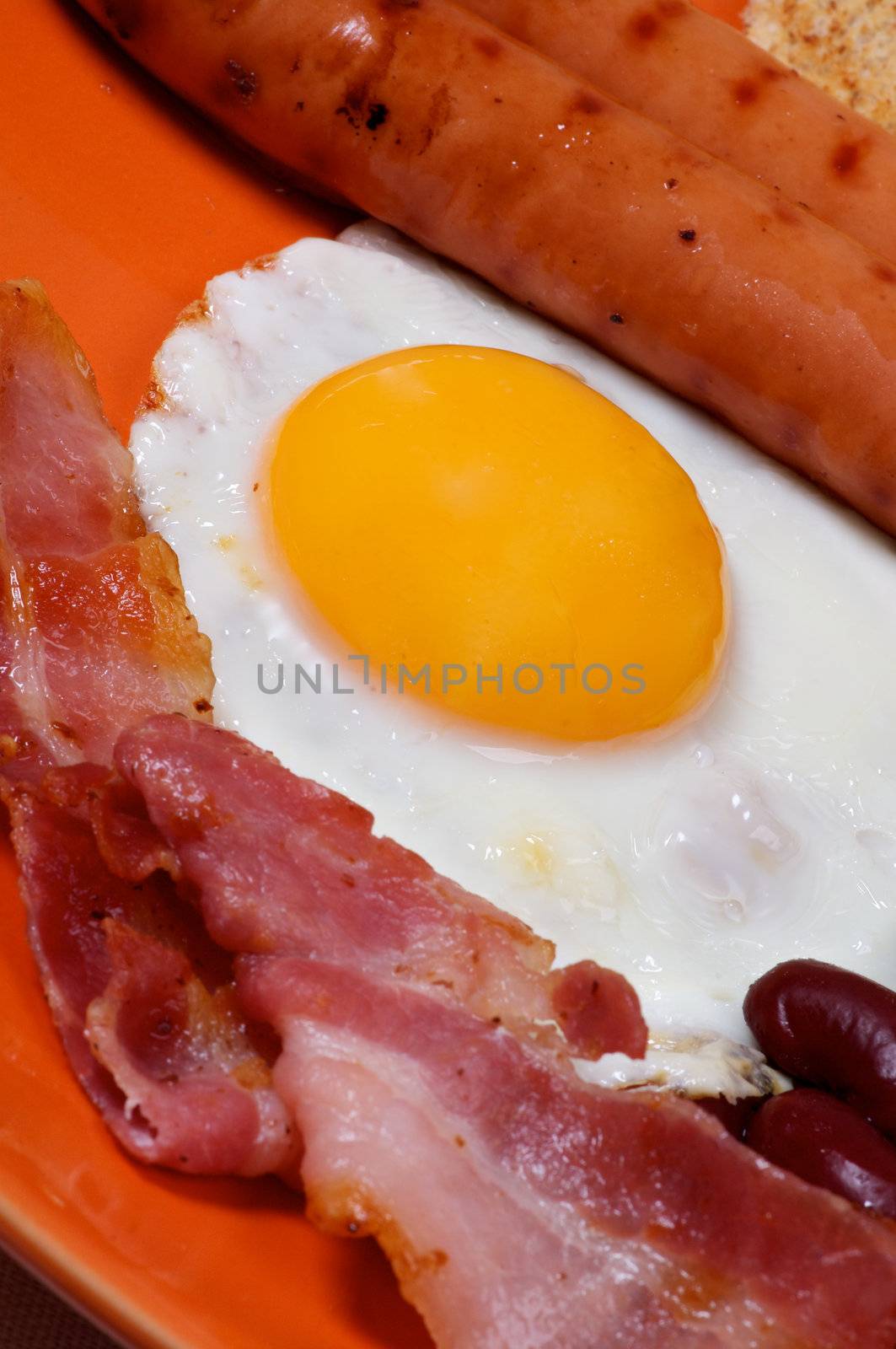 English Breakfast with Sausage, Bacon, Fried Eggs Sunny Side up and  Beans, close up on orange plate. Focus on Egg Yolk