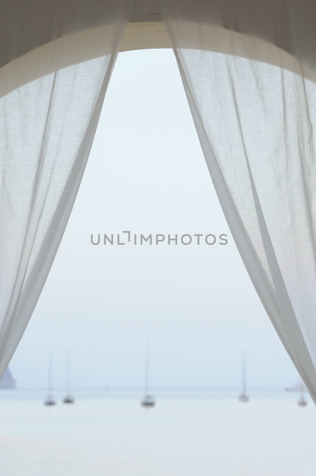 View to distant sailboats by the sea at sunset. Arched balcony with white drapes.