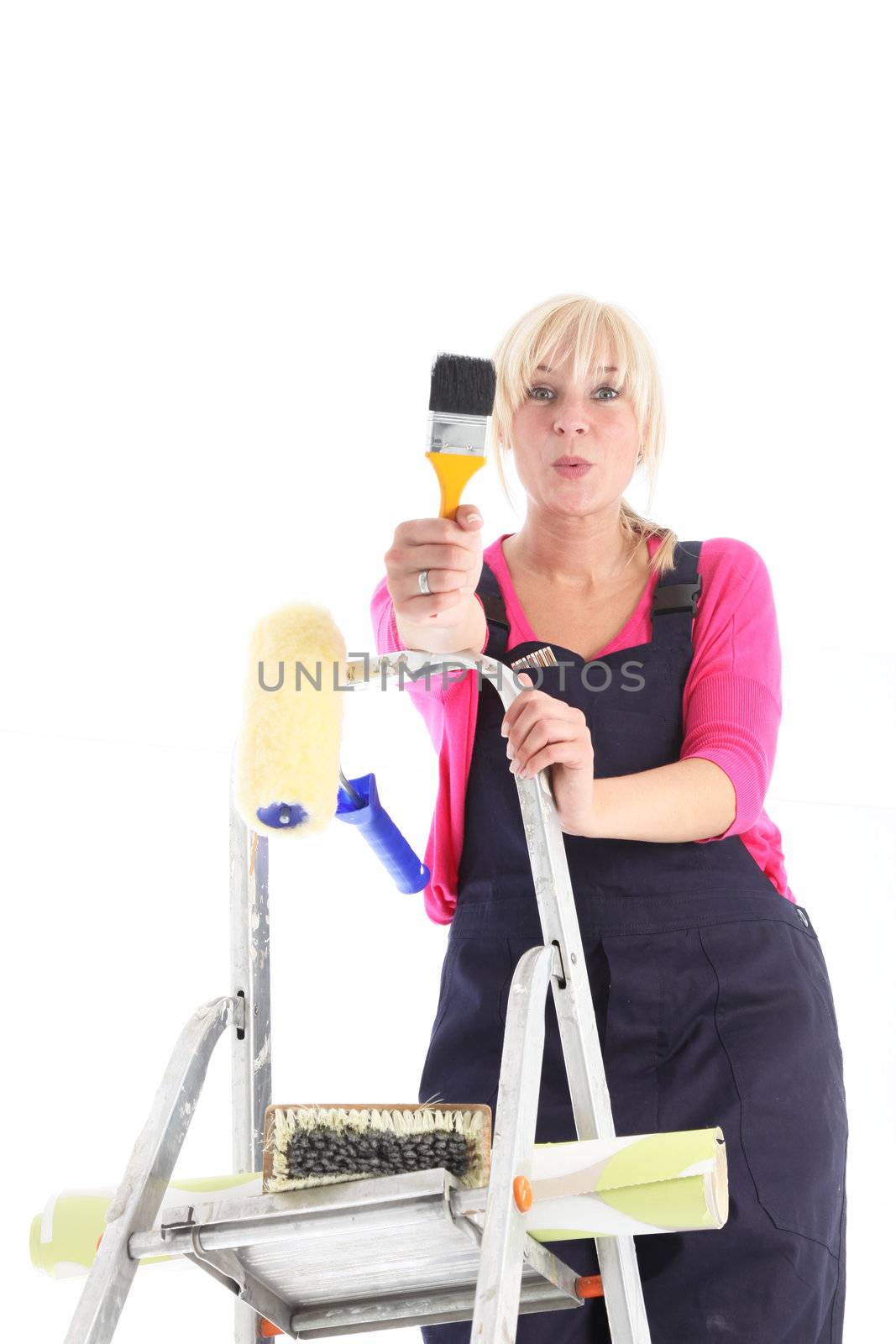 Female decorator wearing dungarees on a ladder doing house redecoration with a roll of wallpaper and brushes isolated on white