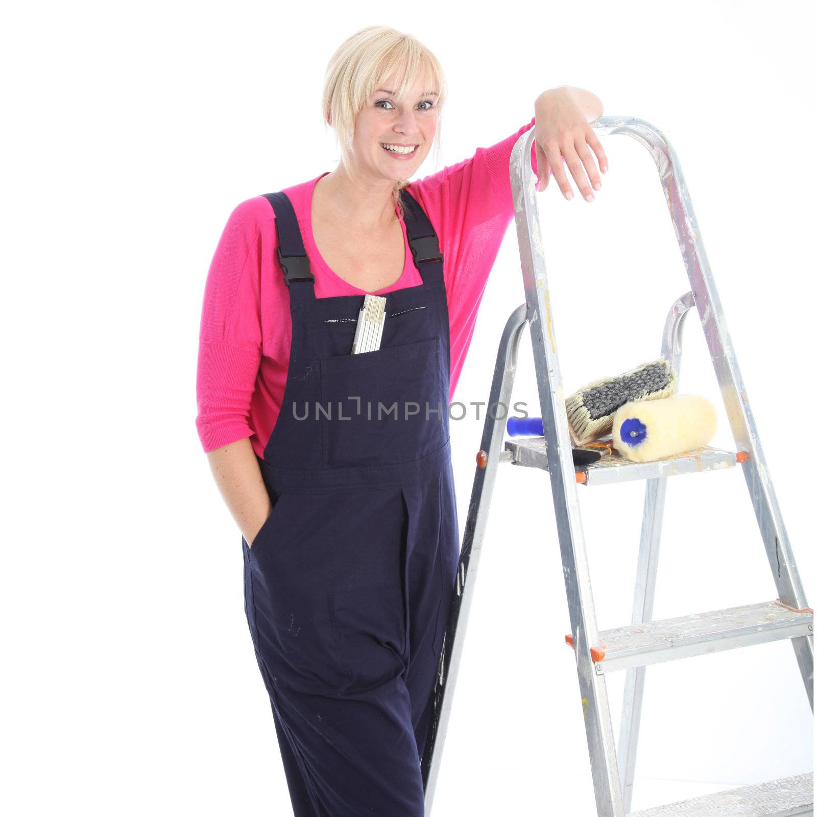 Confident relaxed female decorator standing leaning against a ladder in dungarees with her hand in her pocket isolated on white